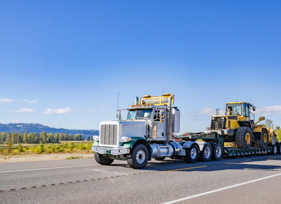 Industrial powerful white day cab big rig semi truck with oversize load sign on the roof transporting heavy oversized tractor on step deck semi trailer running on the highway road