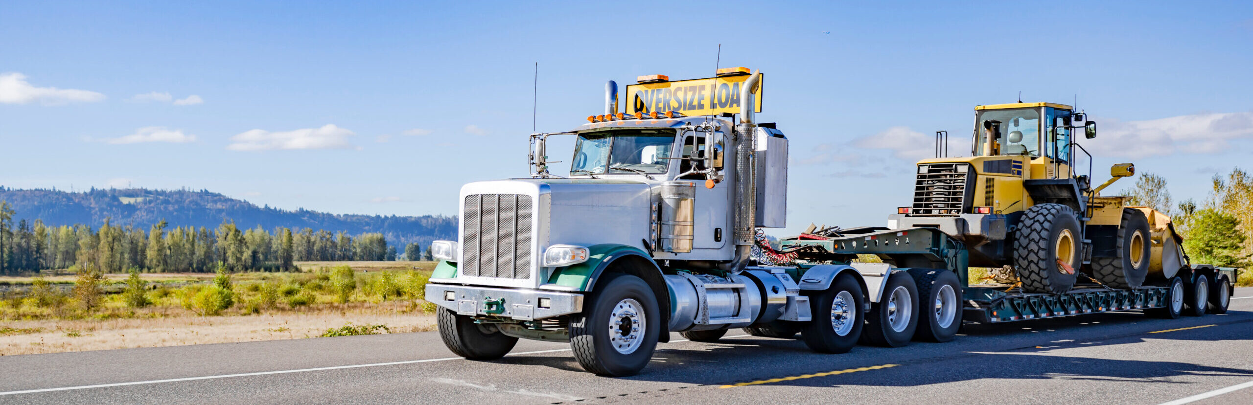 Industrial powerful white day cab big rig semi truck with oversize load sign on the roof transporting heavy oversized tractor on step deck semi trailer running on the highway road