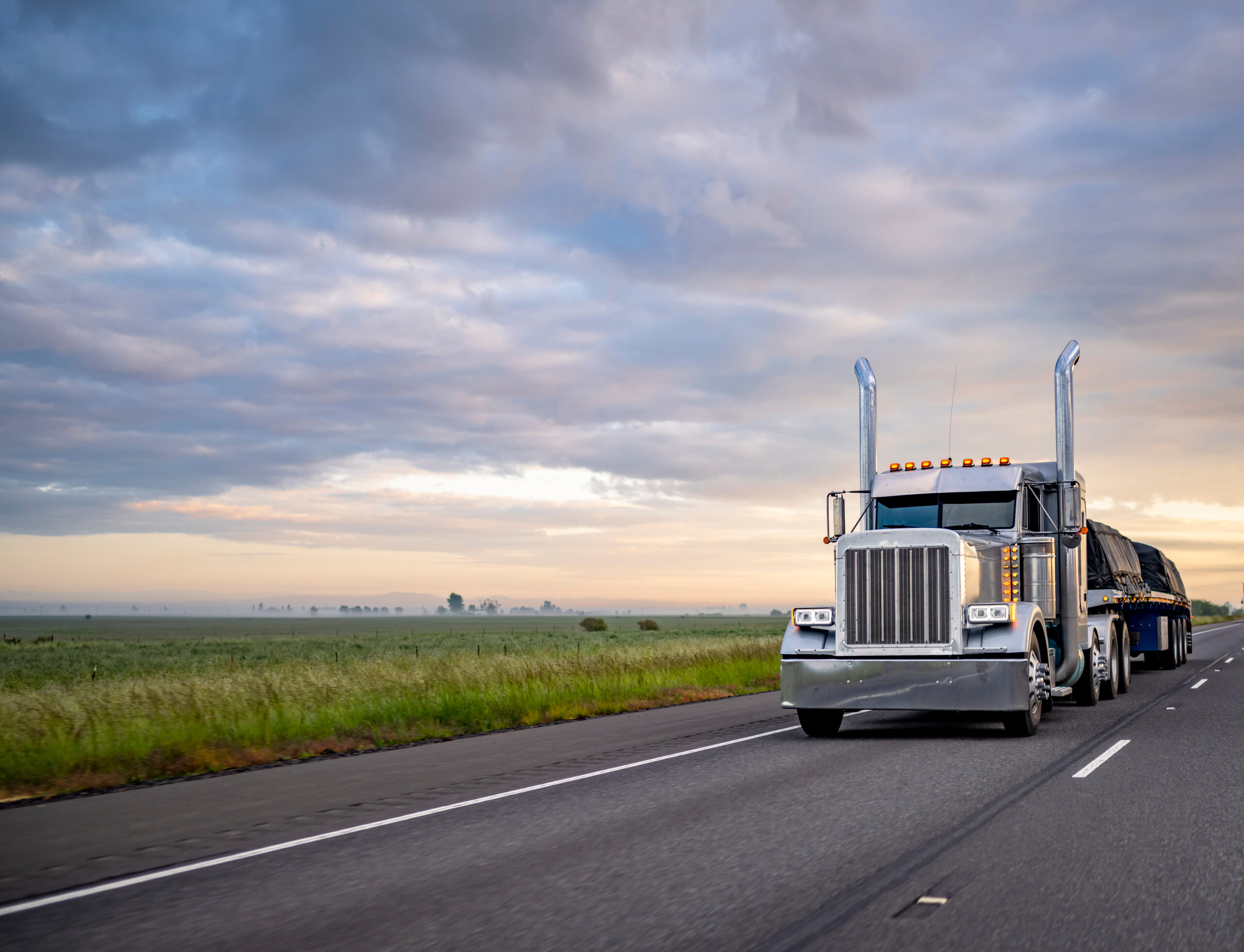 Gray classic big rig American semi truck with turned on lights and chrome exhaust pipes transporting covered cargo on flat bed semi trailer driving on the twilight highway road with stormy clouds sky