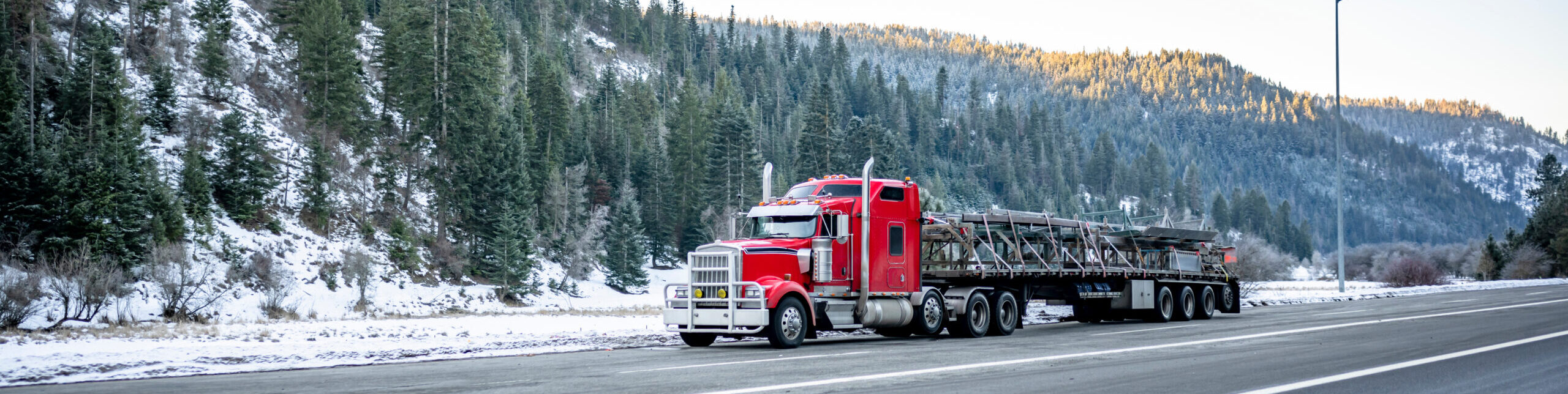 Professional freight red classic big rig semi truck transporting cargo on the long flat bed semi trailer climbing uphill on the winding dangerous slippery winter road with snow and ice in Montana